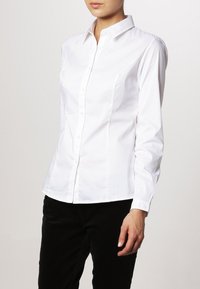 Donna che indossa una camicia bianca aderente con maniche lunghe e colletto, abbinata a pantaloni neri, su uno sfondo uniforme.