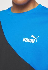 Modré a černé tričko s krátkým rukávem a kulatým výstřihem, které má bílé logo Puma a horizontální design v blokových barvách.
