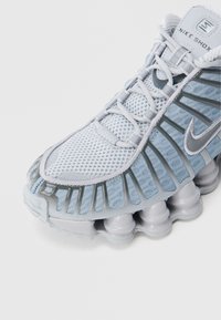 Αθλητικό παπούτσι Nike Shox σε ανοιχτό μπλε και γκρι, με επάνω μέρος από δίχτυ, επενδεδυμένο γιακά, χαρακτηριστικές στήλες απορρόφησης κραδασμών και αντανακλαστικές λεπτομέρειες.