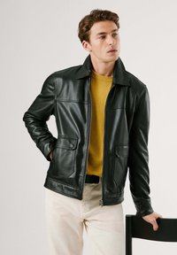 Chaqueta de cuero negra con cremallera frontal, dos bolsillos frontales y cuello con solapa; llevada sobre un suéter amarillo y pantalones de color claro.