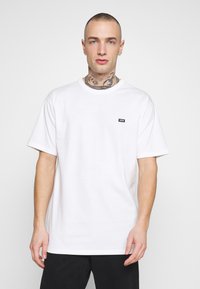 Camiseta blanca de algodón con mangas cortas, un cuello redondo y un pequeño logo negro de Vans en el lado izquierdo del pecho. El portador la presenta contra un fondo claro.