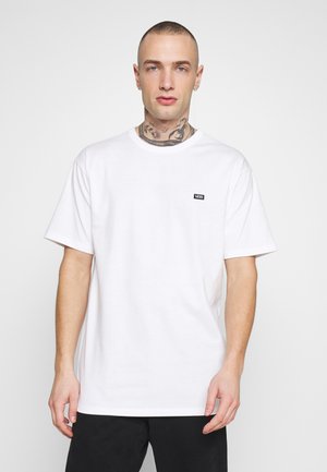 Camiseta blanca de algodón con mangas cortas, un cuello redondo y un pequeño logo negro de Vans en el lado izquierdo del pecho. El portador la presenta contra un fondo claro.