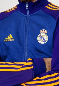 Lila och blå zip-up jacka med gult Adidas-logo och Real Madrid-emblem; har ribbat krage och randiga detaljer på ärmarna.