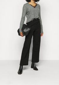 Grijze v-halstrui met kantafwerking, gecombineerd met hoge zwarte wide-leg broek en zwarte enkellaarsjes met hak. Draagt een zwarte tas.