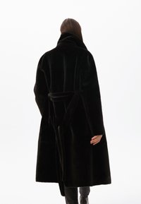Černý dlouhý kabát vyrobený z plyšového materiálu, s širokým límcem, páskem v pase a volným střihem, zobrazený zezadu.