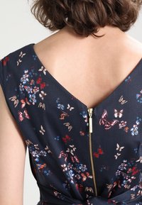 Blommig klänning med mörk marinblå tyg, som har en V-formad ryggkrage, korta ärmar och en synlig gulddragkedja bak.