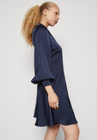 Navy blue satin dress with long puff sleeves and a V-neckline. Features a flared silhouette and subtle button details at the cuffs.