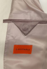 Beige satijnen stof met een driehoekige zak; oranje label met zwarte tekst "SUITABLE" gestikt. Gladde textuur met nette naden.