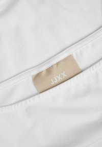 Tela de algodón blanco con una textura suave, que presenta una etiqueta beige con "JJXX" impreso en blanco. La etiqueta está cosida al borde de la tela.