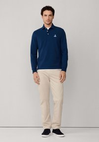 Man staat tegen een eenvoudige achtergrond, gekleed in een marineblauw poloshirt met lange mouwen, beige broek, zwarte sneakers, en een wit T-shirt zichtbaar bij de kraag.