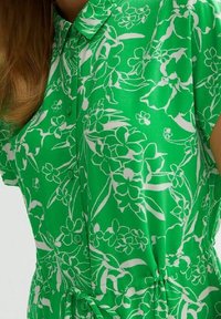 Zelená květinová košile s límečkem na knoflíky, krátkými rukávy a vzorem bílých květin. Měkká látka s detailem stahování v pase na šňůrku.