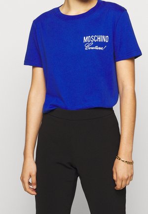 Niebieski bawełniany t-shirt z białym haftowanym napisem "Moschino Couture!" na klatce piersiowej. Krótkie rękawy i okrągły dekolt. Połączony z czarnymi spodniami.