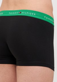 Schwarze Boxershorts mit einem grünen elastischen Bündchen, auf dem "TOMMY HILFIGER" steht. Der Stoff wirkt glatt und eng anliegend. Nahtdetails sind sichtbar.