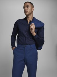 Navy button-up shirt with long sleeves, paired with matching blue dress pants. The shirt has a smooth texture and is tucked in. A blazer rests on the shoulder.