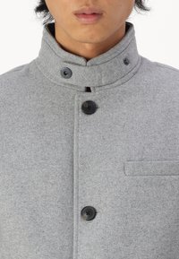 Grå feltjakke med høy krage, med svarte knapper og en brystlomme. Stoffet har en myk tekstur som gir et rent og strukturert utseende.