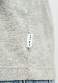 Pilkas medvilninis marškinėlis su minkšta tekstūra, turintis baltą etikėlę šone su užrašu "JACK & JONES". Matoma siuvimo linija apatiniame krašte.