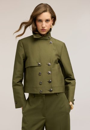 Mujer con chaqueta cruzada de color verde oliva y pantalones a juego, con pendientes de aro dorados y pulsera, de pie contra un fondo liso.