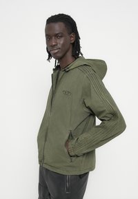 Χακί πράσινο μπουφάν με κουκούλα, με μπροστινό φερμουάρ, πλαϊνές τσέπες και υφασμάτινη ραμμένη λεπτομέρεια κατά μήκος των μανικιών.
