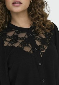 Czarna bluzka z koronkową wstawką w kwiatowe wzory przy yoke, z zapięciem na guziki oraz miękką teksturą materiału.