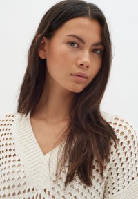 Weißer Strickpullover mit V-Ausschnitt-Design und Lochmuster. Der Stoff wirkt leicht und strukturiert, mit einer lockeren Passform.