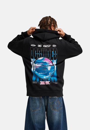 Osoba ubrana w czarną bluzę z kapturem DC Shoes z kolorową grafiką i napisem na plecach, w połączeniu z niebieskimi dżinsami z wyszywanym logo na kieszeni.