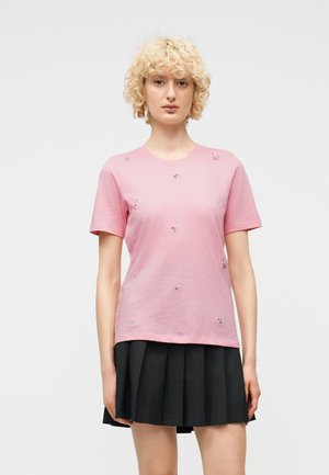 Blond krullend haar persoon die een roze geborduurd shirt met korte mouwen en een zwarte geplooide rok draagt, staand tegen een effen achtergrond.