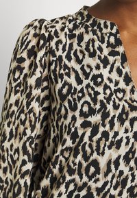 Leopardmönstrad blus med V-ringning, med en krämfärgad bas och bruna samt svarta fläckar, samt strukturerade pufarmar. Mjuk textur och fluffig design.
