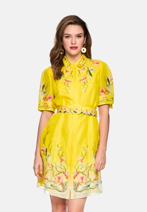 Woman wearing a bright yellow dress with floral embroidery, a matching belt, puffed short sleeves, and large cream-colored earrings.