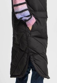 Svart pufferväst med ett quiltat mönster, sidofickor och en hög slits. Bärs över en randig tröja i rosa och lila.