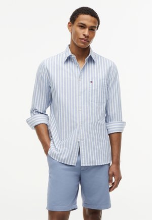 Young man wearing light blue and white vertical striped button-up shirt with rolled sleeves and solid light blue shorts, standing relaxed.