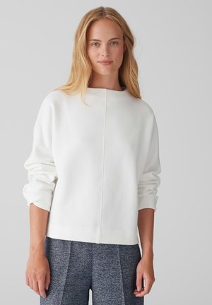 Sweatshirt blanc à col rond, épaules tombantes et poignets côtelés. Texturé lisse, coutures minimales, associé à un pantalon bleu à motifs texturés.