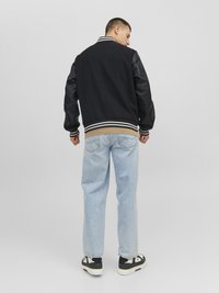 Zwarte varsityjack met leren mouwen, witte strepen op de kraag en manchetten, gecombineerd met lichtblauwe jeans en zwarte sneakers.