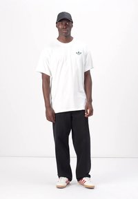 Tricou alb din bumbac cu un logo verde, asortat cu pantaloni negri și adidași crem cu dungi roșii. O șapcă neagră completează ținuta.