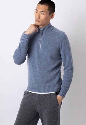 Uomo che indossa un maglione di lana blu con zip, collo e polsini a coste, abbinato a pantaloni grigio scuro, in piedi davanti a uno sfondo chiaro e uniforme.