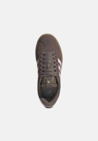 Zapatilla de ante marrón con tres franjas blancas, puntera redondeada, lengüeta acolchada y suela exterior texturizada. Presenta el logo de la marca en la lengüeta y en el lateral.