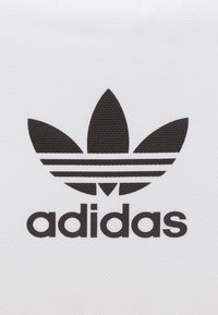 Musta Adidas trefoil -logo, jossa on kolme vaakasuoraa raitaa alhaalla pienellä kirjoitettuna Adidas-teksti valkoisella tekstuuripohjalla.