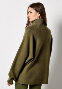 Pull oversized vert olive avec un col haut, des accents côtelés et des détails boutons sur les poignets, fabriqué à partir d'un tissu doux et texturé.