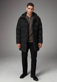 Abrigo de plumas negro con capucha, que cuenta con cierre de cremallera y botones a presión, sobre un suéter de cuello redondo marrón y pantalones negros, combinado con botas negras.