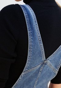 Denim overall with light blue color, adjustable straps, and a crossback design. Textured fabric with visible stitching details.