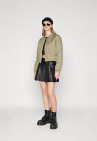 Olivovo zelená bomber jacket s elastickými manžetami, čierny crop top, plisovaná čierna kožená sukňa a čierne šnurovacie topánky s žltými akcentmi.