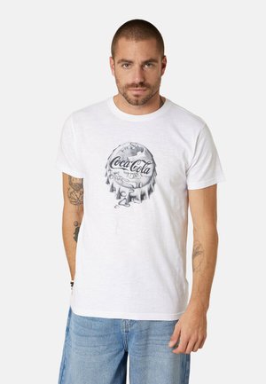 Homme aux cheveux courts et tatouages portant un T-shirt blanc avec un motif de capsule de bouteille Coca-Cola grise et un jean bleu, debout devant un fond blanc.