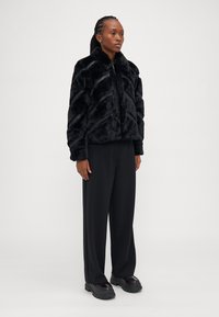 Μαύρο ιαπωνικό μπουφάν από ψεύτικη γούνα με ραβδωτή υφή, διαθέτει ψηλό γιακά και κλείσιμο με φερμουάρ. Συνδυάζεται με φαρδιά μαύρα παν trousers και χοντρά μαύρα υποδήματα.