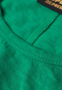 Groen katoenen stof met een gladde textuur, voorzien van een ronde halslijn en doorgestikte randen, met een subtiel gemêleerd patroon.