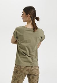 Olivgrünes, kurzärmliges T-Shirt mit lockerer Passform und umgeschlagenen Ärmeln, kombiniert mit beigen Hosen mit floralen Mustern und Taschen auf der Rückseite.