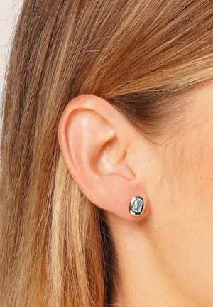 Ohr einer Frau mit glattem, hellbraunem Haar und einem kleinen silbernen Stecker mit einem rechteckigen hellblauen Edelstein.