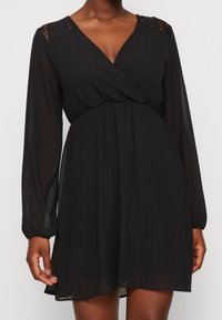 Robe noire en tissu léger avec un col en V, des manches longues et des accents en dentelle sur les épaules. Coupe fluide avec une taille cintrée.