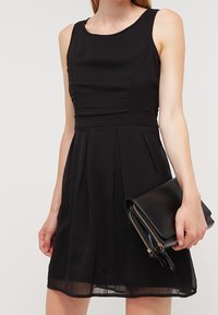 Černé bezramínkové šaty s těsným korzetem, plisovanou sukní a strukturovanou tkaninou; doplněné černou psaníčkem se zlatými zipovými detaily.