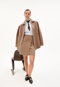 Camicia overshirt in misto lana marrone con grandi tasche, camicia bianca e gonna marrone. Ballerine nere completano l'outfit. Design semplice, colori neutri.