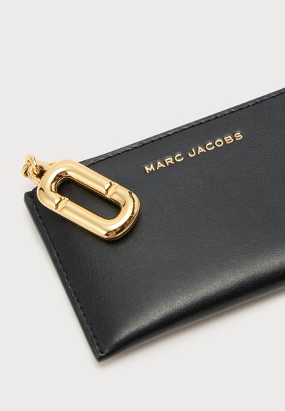 Pochette en cuir noir Marc Jacobs avec texte du logo doré et grande fermeture ovale dorée sur fond clair.