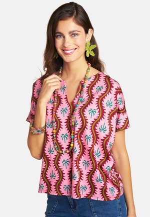 Woman with long brown hair wearing a pink patterned shirt, colorful beaded necklace and bracelet, and green leaf-shaped earrings smiling.